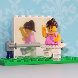 May include: A Lego ballerina figure in a pink leotard and white tutu, with a brown bun, stands on a green base. A mirror reflects her image, and a ballet barre is in front of the mirror. The background is a light blue wall with a floral pattern.