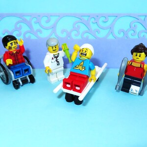 May include: A group of Lego figures depicting a medical scene. One figure is in a wheelchair, another is on a stretcher, and a third is using crutches. A doctor figure is also present, holding a green syringe. The figures are on a blue surface.