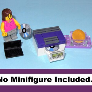 May include: A Lego set featuring a minifigure, a purple and white CD player, a black laptop, and a gold disc on a clear stand. The set includes a CD and a digital display on the CD player. The text "No Minifigure Included" is displayed.