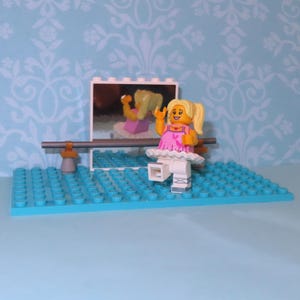 May include: A Lego ballerina figure in a pink dress and white tutu, with blonde hair, is positioned on a blue base. A mirror reflects the figure. A ballet barre is in front of the mirror. The background is a light blue wall with a white floral pattern.