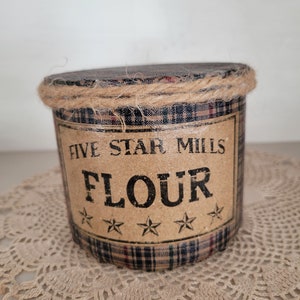 May include: A brown and black plaid canister with a burlap lid and a label that reads "Five Star Mills Flour" with three stars underneath the word "Flour".