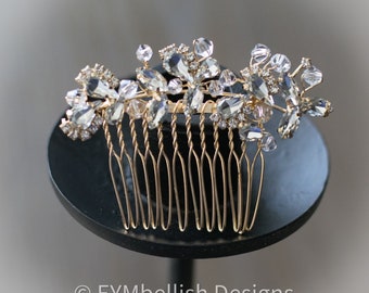 May include: A gold-toned hair comb with clear crystal accents. The comb features a floral design with various sizes of crystals and a row of gold-colored teeth. The comb is displayed on a black stand.