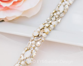 May include: A close-up of a bridal belt with white and clear crystals, pearls, and gold accents. The belt is on a white surface with a pink flower in the background. The belt is embellished with various shapes and sizes of crystals.