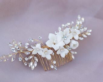 May include: A gold hair comb with white ceramic flowers and clear rhinestones. The comb is decorated with a floral design and is perfect for a wedding or special occasion.