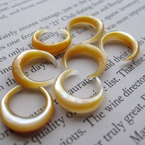 May include: Several iridescent, crescent-shaped ear gauges in shades of yellow and cream are arranged on a page of printed text. The gauges have a smooth, polished surface and a curved design.