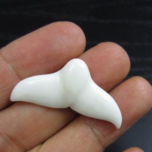 May include: A small, white, whale tail figurine. The tail is smooth and curved, with a rounded top and two flukes. The figurine is held in a person's hand, with the background out of focus.
