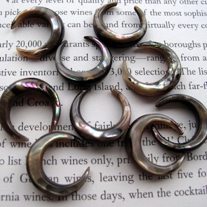 May include: A collection of iridescent, crescent-shaped ear gauges. These dark, semi-circular plugs are made of a material that reflects multiple colors. The gauges are displayed on a background of printed text.