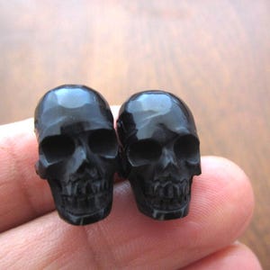May include: Two small, black skull figurines with detailed facial features. The skulls are glossy and appear to be made of a smooth, dark material. The image is a close-up, highlighting the intricate carvings and the overall gothic theme.