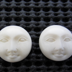 Könnte beinhalten: Zwei runde, cremefarbene Cabochons in Gesichtsform mit geschnitzten Merkmalen, darunter Augen, Nase und lächelnder Mund. Die Cabochons sind glatt und leicht glänzend. Der Hintergrund ist eine strukturierte schwarze Oberfläche.