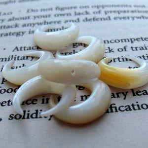 May include: A collection of off-white, curved ear gauges and plugs. The jewelry pieces are made of a smooth, glossy material, with a few showing a hint of yellow. They are scattered on a page of printed text.