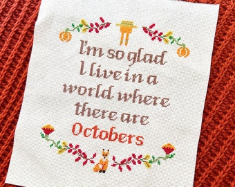 May include: Cross-stitch embroidery on white fabric with a fall-themed quote: "I'm so glad I live in a world where there are Octobers." The design includes a scarecrow, pumpkins, leaves, and foxes.