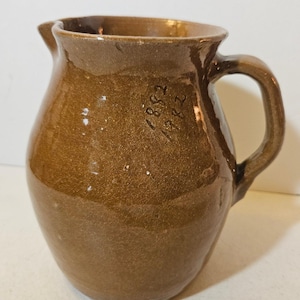 May include: A brown glazed ceramic pitcher with a rounded body, a curved handle, and a pouring spout. The surface features a glossy finish with visible crazing and the hand-etched numbers 1882 and 1982 inscribed on the upper side of the vessel.