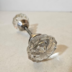 May include: A clear glass door knob set featuring two fluted, circular glass handles connected by a central metal spindle. Each glass knob has a scalloped edge and is attached to a polished silver-toned metal base that secures the glass to the rod.