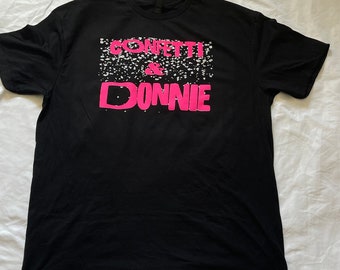 Könnte beinhalten: Schwarzes T-Shirt mit einem rosafarbenen Grafikdruck, auf dem "CONFETTI & DONNIE" steht, mit weißen Konfetti, die um den Text herum verstreut sind.