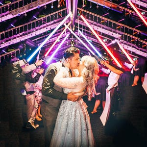 May include: A couple kissing under an arch of glowing blue and red light sabres. The person on the left wears a dark waistcoat and shirt, while the person on the right wears a long, shimmering silver gown with star patterns, surrounded by guests.