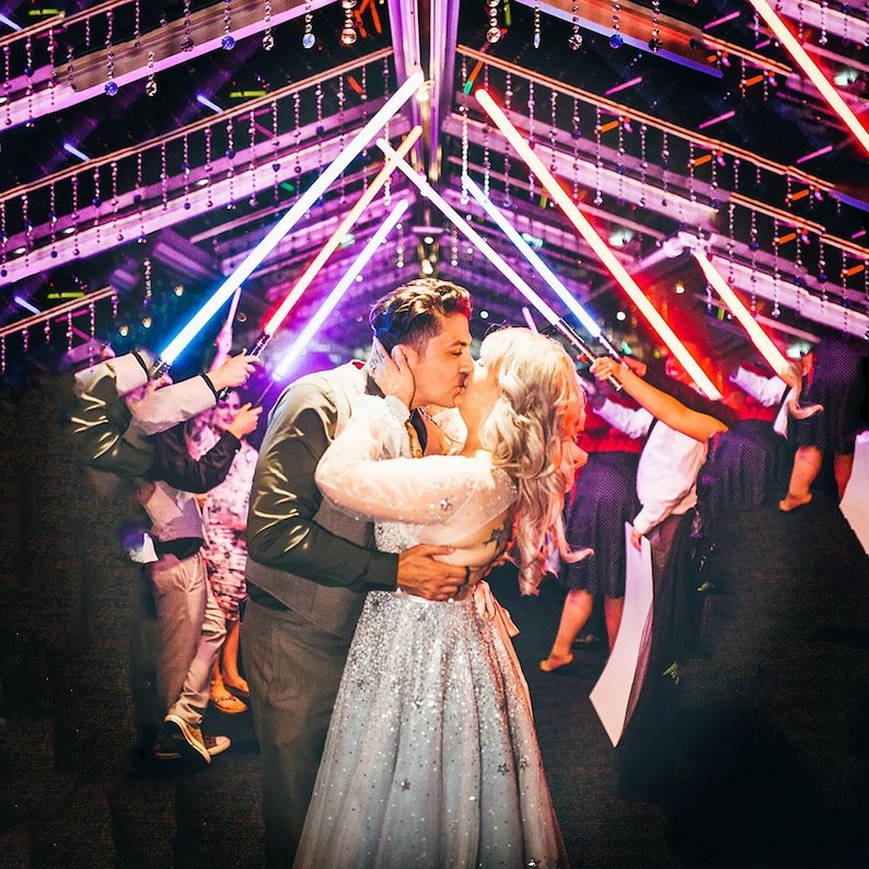 May include: A couple kissing under an arch of glowing blue and red light sabres. The person on the left wears a dark waistcoat and shirt, while the person on the right wears a long, shimmering silver gown with star patterns, surrounded by guests.