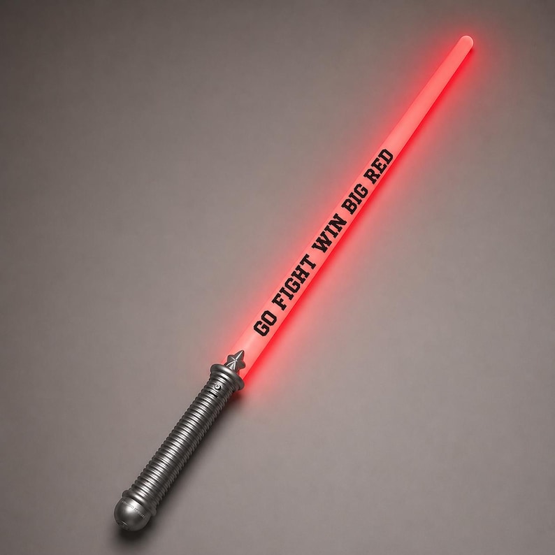 May include: A light-up toy sword with a silver-coloured handle and a glowing red blade. The blade features the black text "GO FIGHT WIN BIG RED" printed vertically along its length. The handle has a ribbed texture and a decorative pommel design.