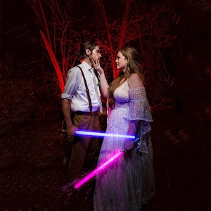May include: A white button-down shirt with brown braces and tan trousers paired with a white lace off-the-shoulder gown. Both individuals hold glowing light-up swords, one emitting a bright blue light and the other a vibrant pink light.
