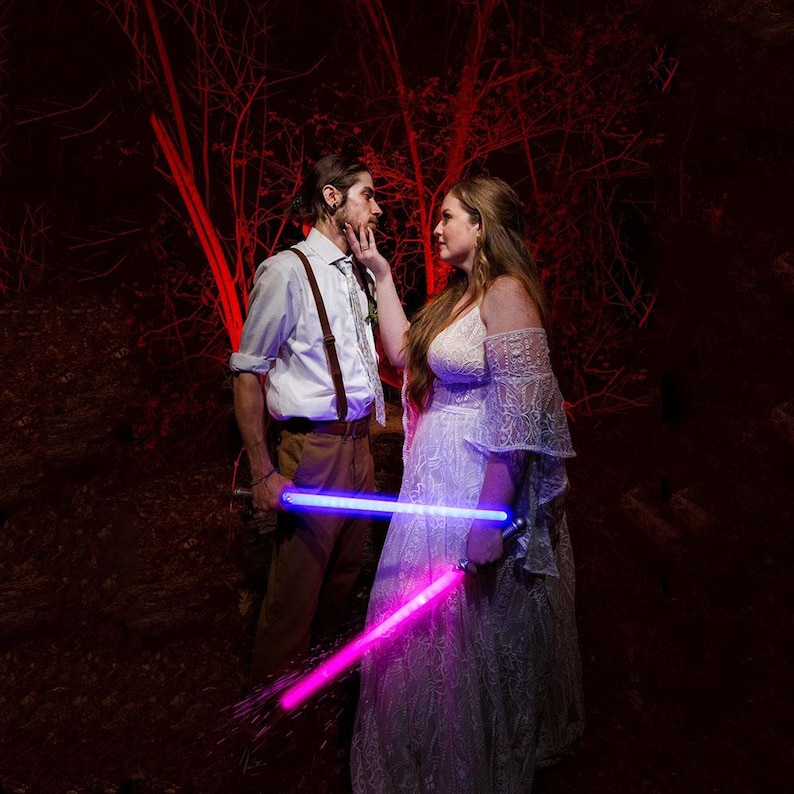 May include: A white button-down shirt with brown braces and tan trousers paired with a white lace off-the-shoulder gown. Both individuals hold glowing light-up swords, one emitting a bright blue light and the other a vibrant pink light.