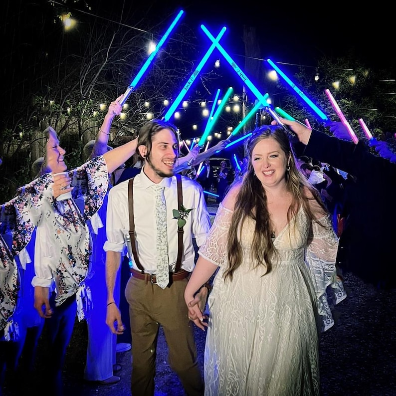 May include: A couple walking through a tunnel of glowing blue and pink light-up swords held by guests. The person on the left wears a white shirt, brown braces, and a patterned tie. The person on the right wears a white lace dress with sleeves.