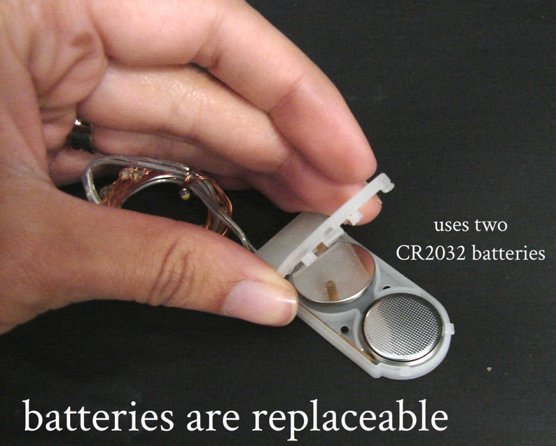 K&ouml;nnte beinhalten: Ein wei&szlig;es Batteriefach aus Kunststoff mit einem durchsichtigen Deckel, der offen ist und zwei silberne CR2032-Batterien zeigt. Der Text "verwendet zwei CR2032-Batterien" ist unter dem Fach sichtbar. Der Text "Batterien sind austauschbar" ist unter dem Fach sichtbar.