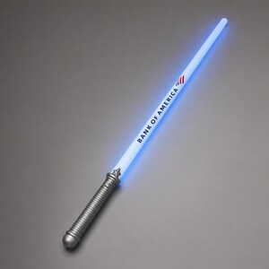 May include: A light-up toy sword with a silver-coloured ribbed handle and a glowing blue blade. The blade features the text BANK OF AMERICA printed in black along with the company's red, white, and blue flag logo near the top of the handle.