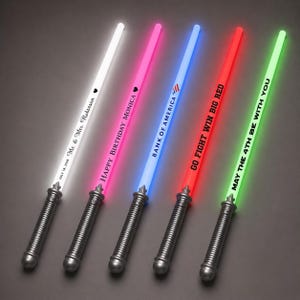 May include: Five light-up toy swords with grey handles and glowing blades in white, pink, blue, red, and green. The blades feature custom text including "Mr. & Mrs. Robinson", "HAPPY BIRTHDAY MONICA", "BANK OF AMERICA", "GO FIGHT WIN BIG RED", and "MAY THE 4TH BE WITH YOU".