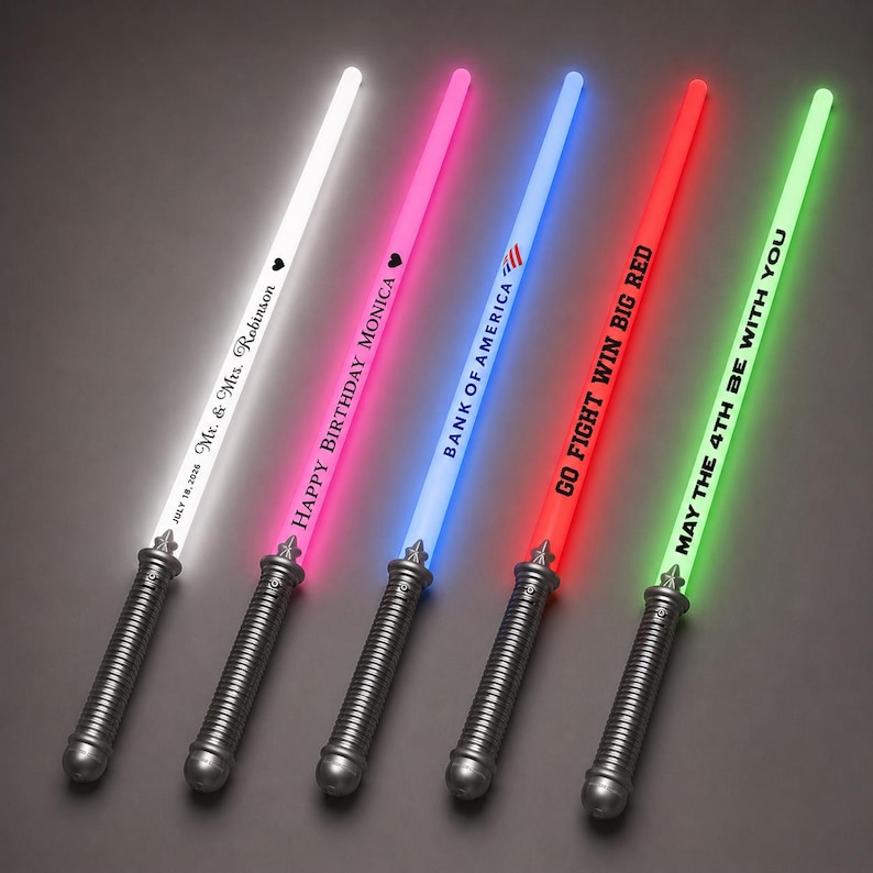 May include: Five light-up toy swords with grey handles and glowing blades in white, pink, blue, red, and green. The blades feature custom text including "Mr. & Mrs. Robinson", "HAPPY BIRTHDAY MONICA", "BANK OF AMERICA", "GO FIGHT WIN BIG RED", and "MAY THE 4TH BE WITH YOU".