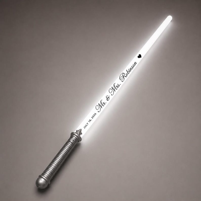 May include: A glowing white light sabre prop with a silver metallic handle. The blade features the black text "Mr. & Mrs. Robinson" followed by a small heart icon and the date "JULY 18, 2026" printed vertically along the length of the illuminated shaft.