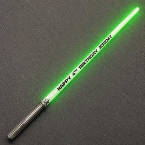 May include: A glowing green toy sword with a metallic-coloured ribbed handle. The blade features the black printed text HAPPY 4TH BIRTHDAY JEREMY. The sword is positioned diagonally across the frame, emitting a bright light from the long blade.