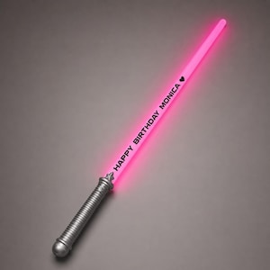 May include: A glowing pink light-up toy sword with a silver-coloured handle. The blade features the black text "HAPPY BIRTHDAY MONICA" followed by a small black heart symbol. The toy sword is positioned diagonally against a neutral grey background.
