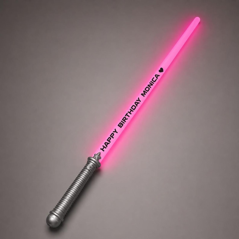 May include: A glowing pink light-up toy sword with a silver-coloured handle. The blade features the black text "HAPPY BIRTHDAY MONICA" followed by a small black heart symbol. The toy sword is positioned diagonally against a neutral grey background.
