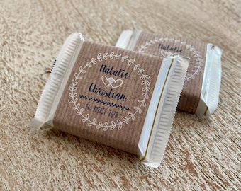 Pode incluir: Dois pequenos favores embrulhados em papel castanho com guarnições brancas e detalhes dourados. Os favores têm uma coroa branca com o texto "Natalie Christian" e "04. August 2020" impresso em preto.