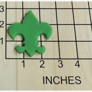 May include: A green plastic fleur-de-lis cookie cutter, approximately 1.5 inches wide and 1.5 inches tall, on a wooden surface with a grid pattern.
