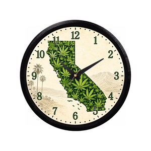 May include: A round wall clock with a black frame and a cream-colored face. The center features a map of California filled with a green cannabis leaf pattern. The background shows a landscape with palm trees and mountains. Black hands indicate time.
