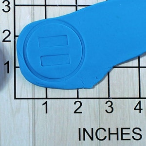 May include: A blue silicone mold with a circular design featuring two rectangular cutouts. The mold is approximately 2 inches wide and 1 inch tall.