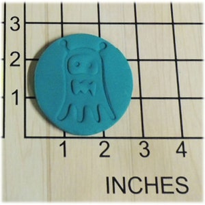 May include: A teal blue round cookie cutter with a design of a cartoon alien wearing a bow tie. The cookie cutter is approximately 2 inches in diameter.