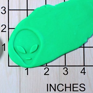 May include: A green alien face cookie cutter, measuring approximately 2 inches in length, on a wooden surface with a ruler.