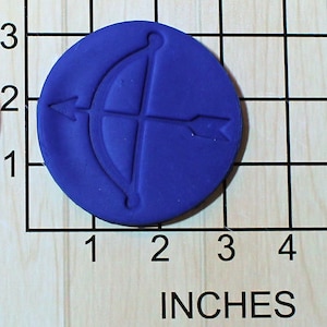May include: A round, blue, polymer clay stamp with a raised design of a bow and arrow. The stamp is approximately 2 inches in diameter.