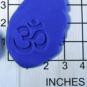 May include: A close-up of a blue silicone mold with an Om symbol embossed in the center. The mold is approximately 2 inches wide and is sitting on a gridded surface.