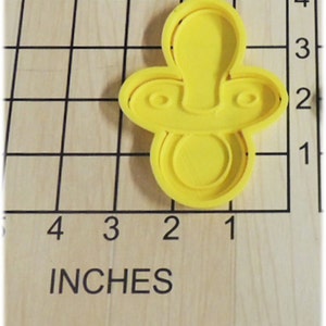 May include: A yellow plastic pacifier-shaped cookie cutter. The cutter is approximately 2 inches long and 1.5 inches wide. The cutter is on a wooden surface with a grid and the word "INCHES" printed on it.