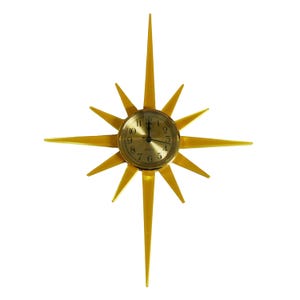 May include: A gold-colored starburst wall clock with a round clock face. The clock has a gold metal frame with pointed rays extending outward. The clock face has black numerals and hands.