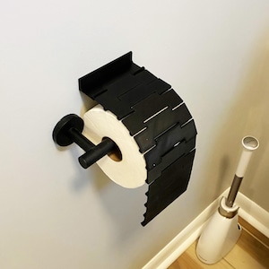 May include: A black, geometric toilet paper holder mounted on a light gray wall. The holder has a unique, segmented design and holds a roll of white toilet paper. A matching black toilet paper holder and a white and silver toilet brush are also visible.
