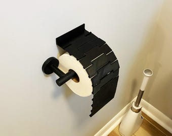 May include: A black, geometric toilet paper holder mounted on a light gray wall. The holder has a unique, segmented design and holds a roll of white toilet paper. A matching black toilet paper holder and a white and silver toilet brush are also visible.