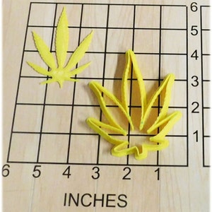 May include: Two yellow cannabis leaf-shaped items on a wooden surface with a grid and the word "INCHES" printed at the bottom. One leaf is a solid shape, and the other is a cookie cutter. The grid shows measurements in inches.