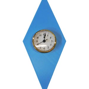 May include: A wall clock with a gold-colored bezel and a white face, mounted on a blue diamond-shaped background. The clock hands point to approximately 1 o'clock. The background is a solid, light blue color.