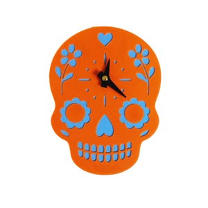 May include: Orange skull-shaped wall clock with blue floral and heart accents. The clock features black hands and a decorative design, perfect for adding a unique touch to any room. The clock's design is reminiscent of the Day of the Dead.