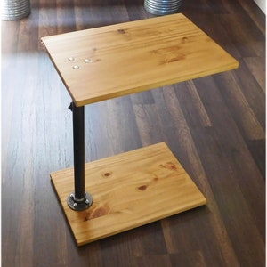 May include: A wooden side table with a black metal pipe leg. The table has a rectangular top and a smaller rectangular shelf underneath. The table is made of natural wood with a light brown finish.