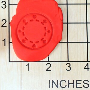 May include: A red clay impression of a poker chip with a circle of hearts around the edge. The clay is on a wooden surface with a grid pattern.