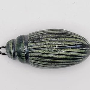 Può includere: Un ciondolo a forma di scarabeo verde e nero con una superficie strutturata. Il ciondolo ha un anello nella parte superiore per essere appeso ed è disegnato con strisce orizzontali. La tavolozza dei colori include sfumature di verde e nero.